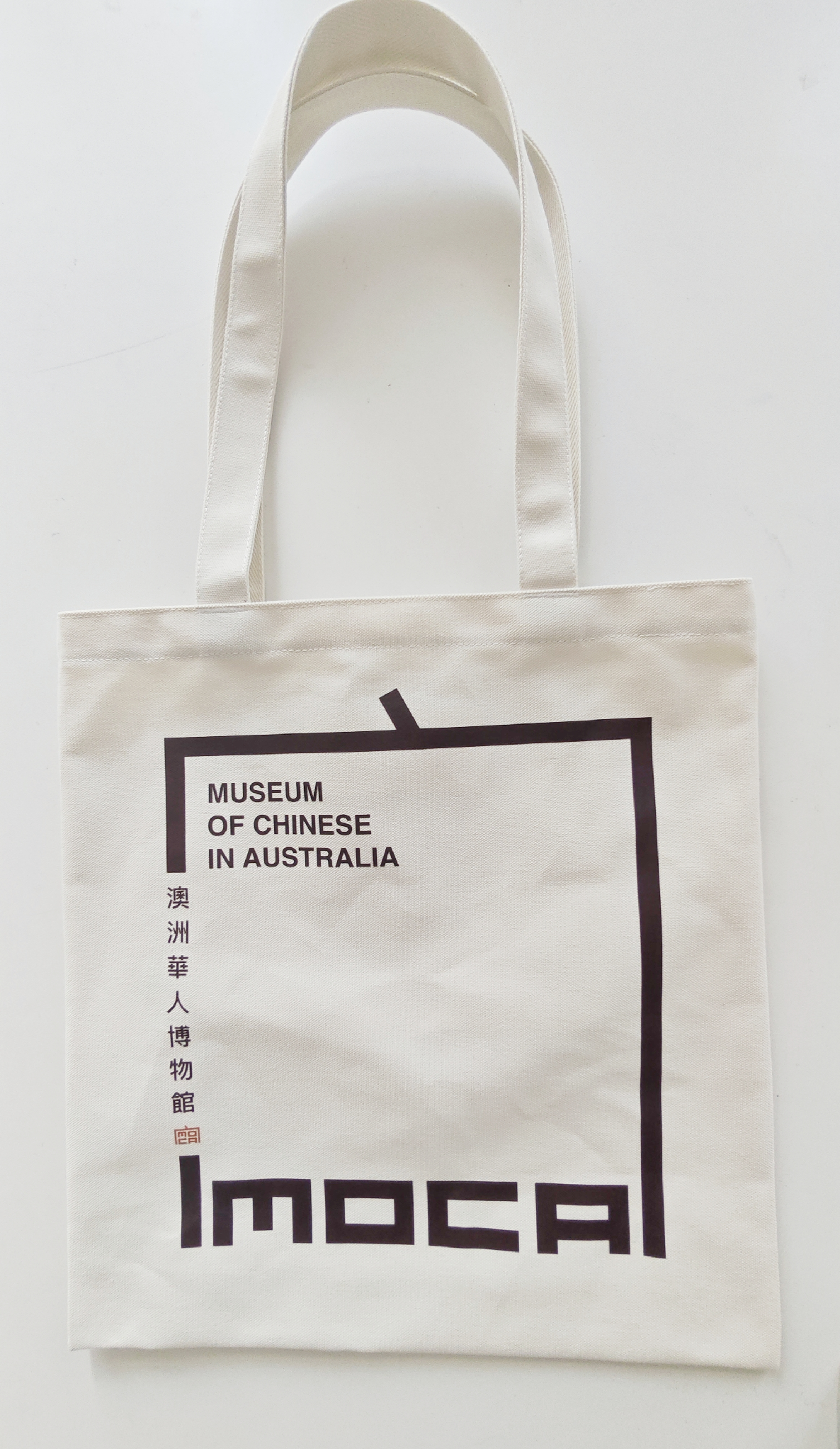Reverse of a natural-coloured tote bag with the Museum of Chinese in Australia bilingual identity in dark brown.