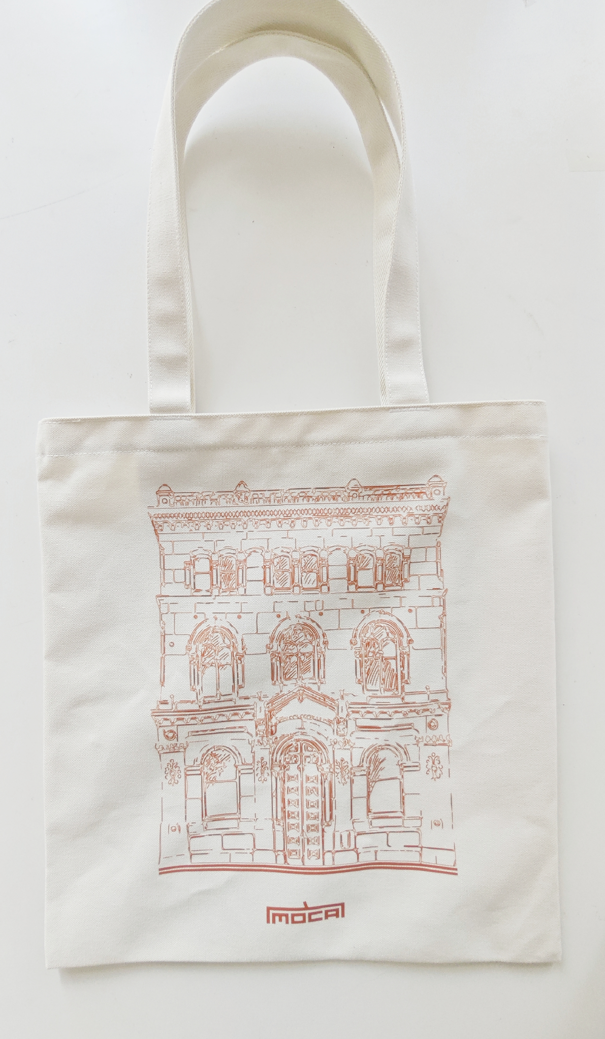 Natural-coloured tote bag with a red line drawing of MOCA's heritage building and the red MOCA wordmark.