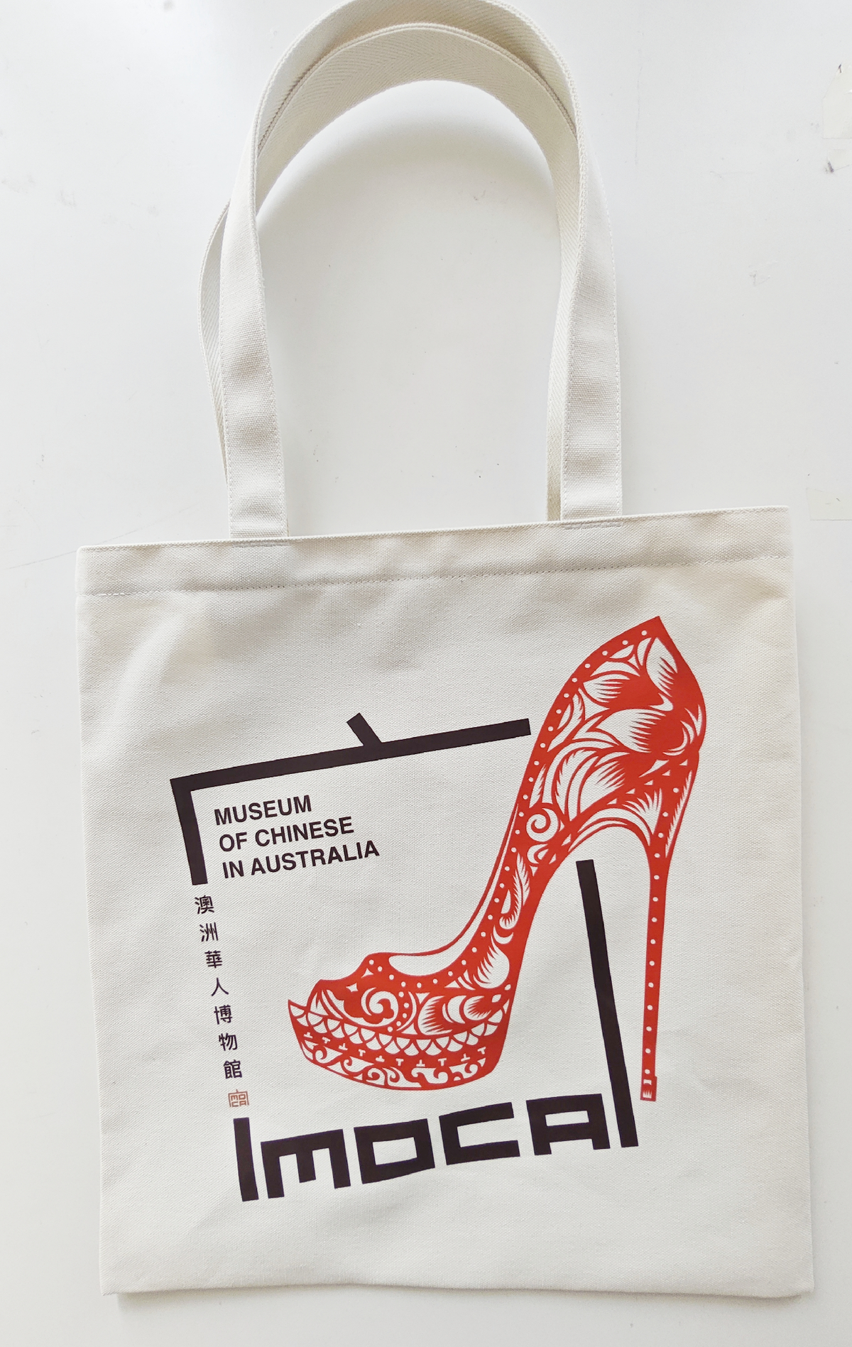 Natural-coloured tote bag with a large red papercut high heel beside the Museum of Chinese in Australia identity.